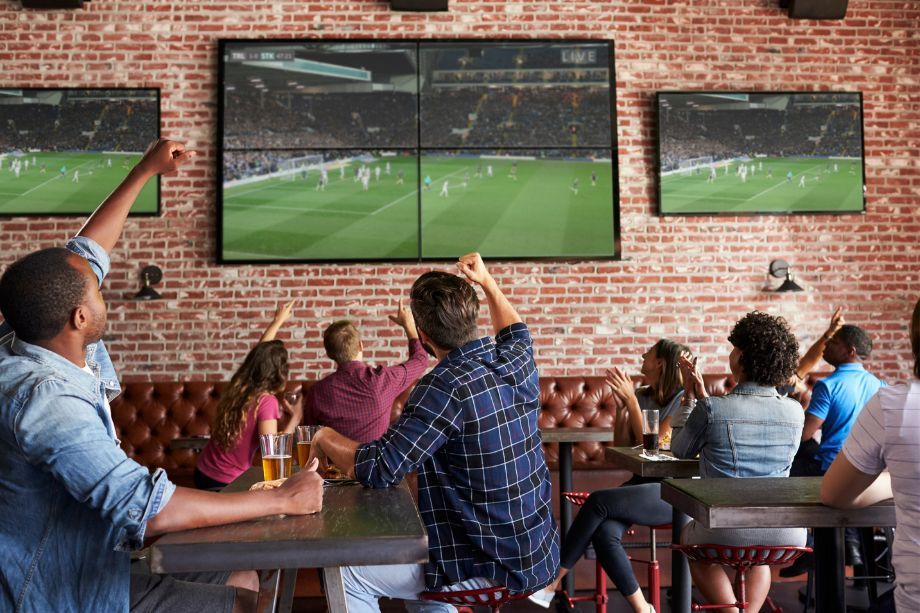 Group of friends in a brick-walled sports bar cheering while watching a soccer match on multiple wall TVs from a long table.