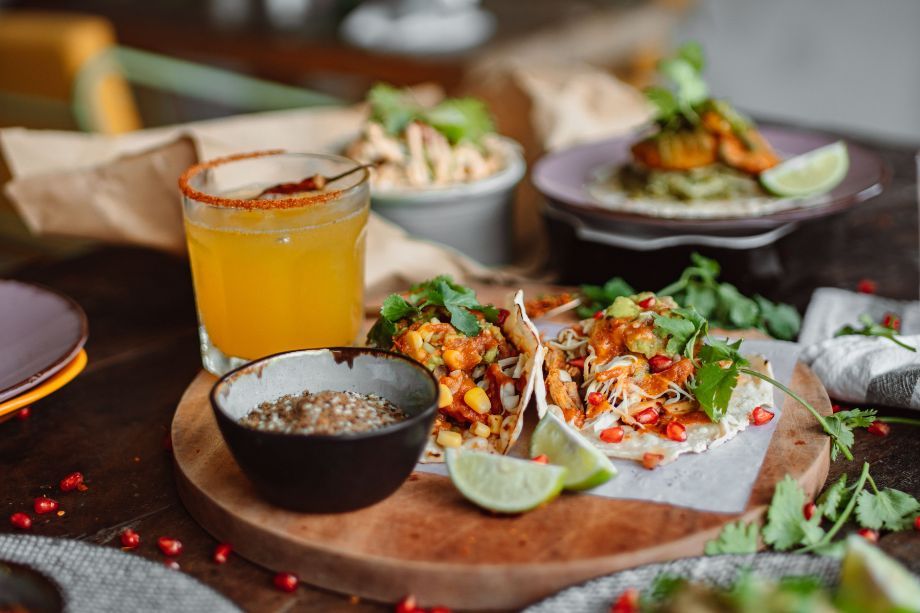 Two tacos on a wooden board topped with cilantro, onions, and peppers, with lime wedges and a side bowl of salsa; a salted drink behind them.