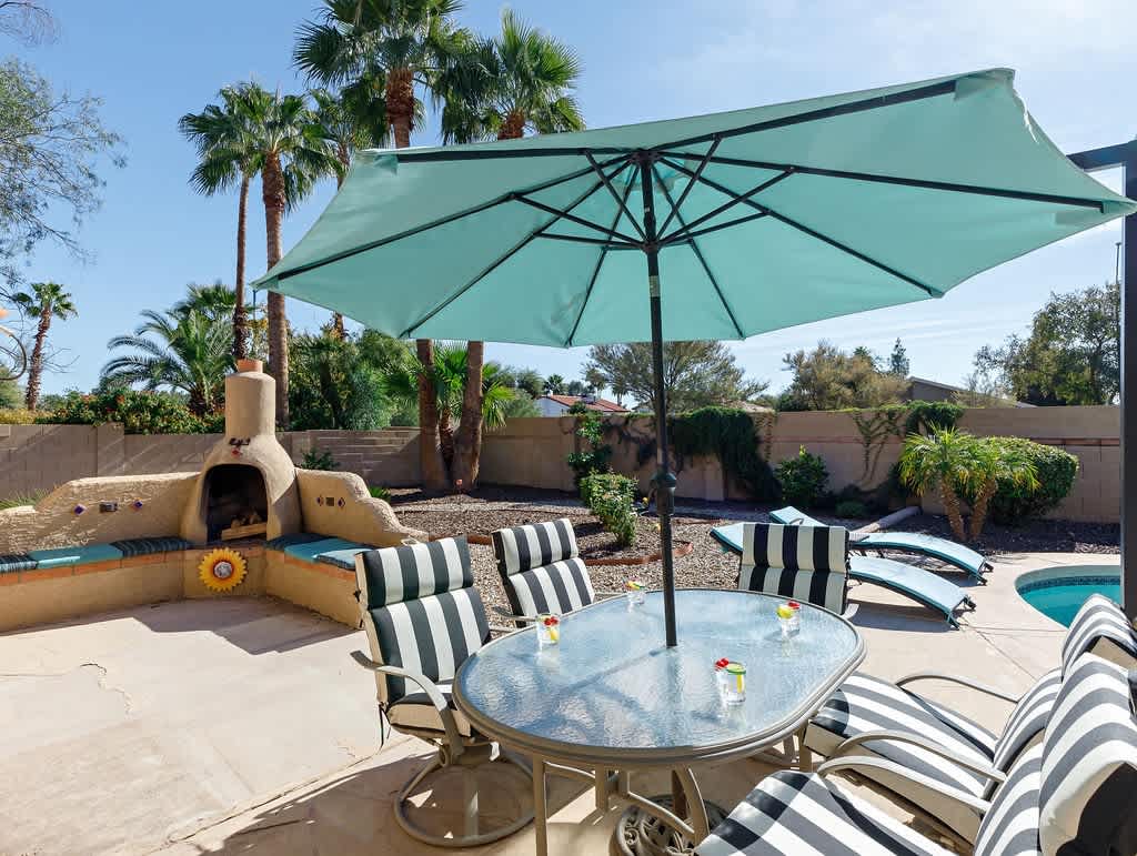 Large private backyard with a sparkling pool, diving board, covered patio with outdoor dining area, sun loungers, and outdoor fire place.