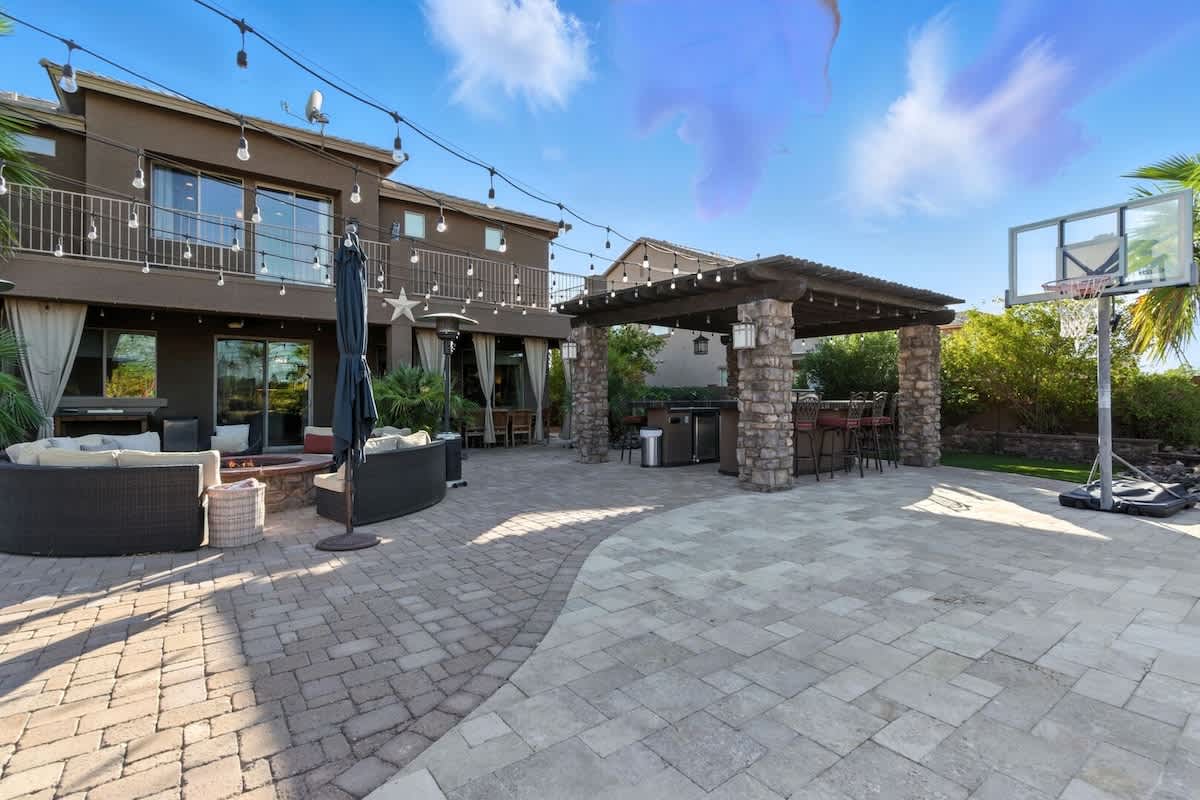 Outdoor fire pit with lounge areas, umbrellas, and outdoor kitchen with BBQ grill, griddle, small refrigirator, and bar stool seating.