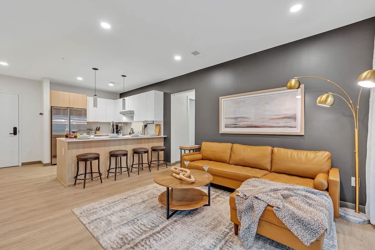 Modern living room decorated in calming tones, featuring comfortable sectional and flat-screen smart TV, and is connected to the kitchen.