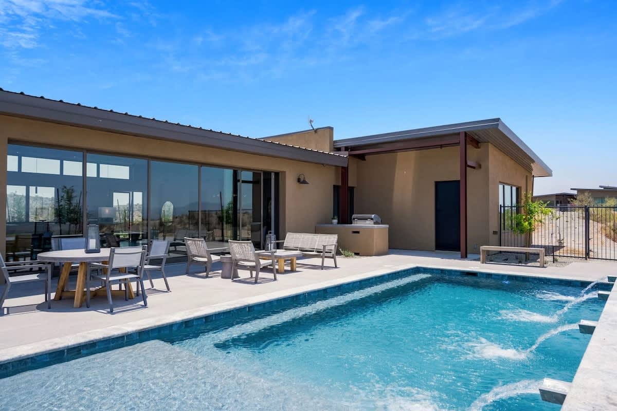 Stunning private oasis featuring a sparkling blue pool with sun loungers, a patio with a BBQ grill, outdoor furnishings for lounging and dining, and a fire pit surrounded by unparalleled desert views.