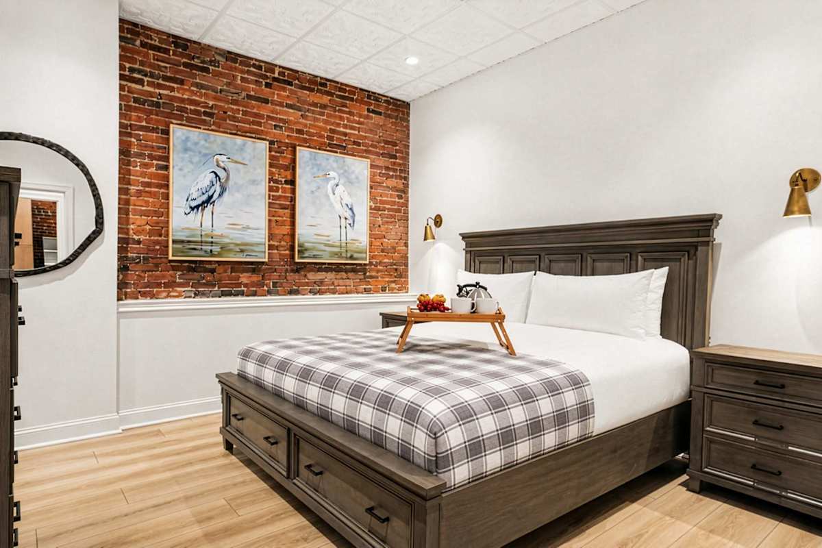 This cozy primary suite features an exposed brick accent wall, plush king bed, and mounted TV, all with an ensuite bath just steps away.