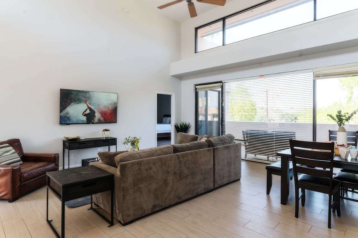 Bright and open living room with soaring ceilings, oversized windows, a plush sectional, Smart TV, and easy flow to the dining area, kitchen, and balcony.