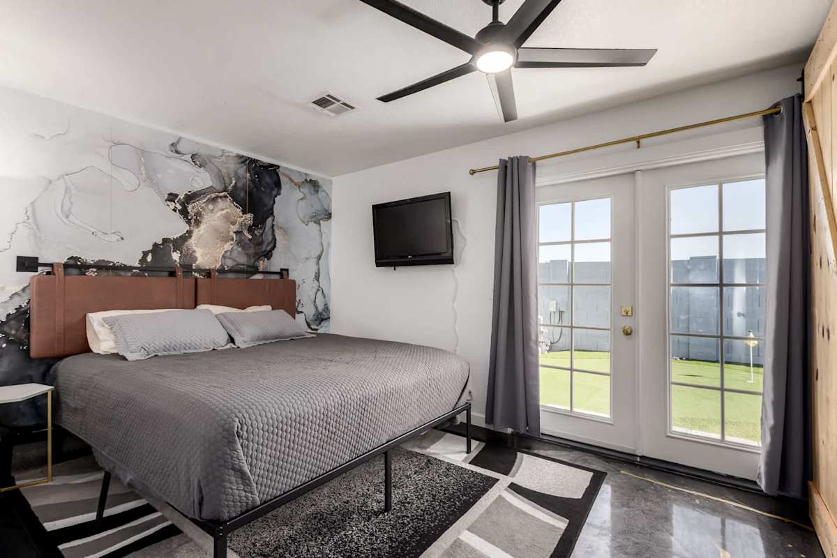 The primary suite offers a king bed, wall-mounted TV, and an en-suite bathroom, with private glass doors opening to backyard access and a moody marble mural adding a modern edge.