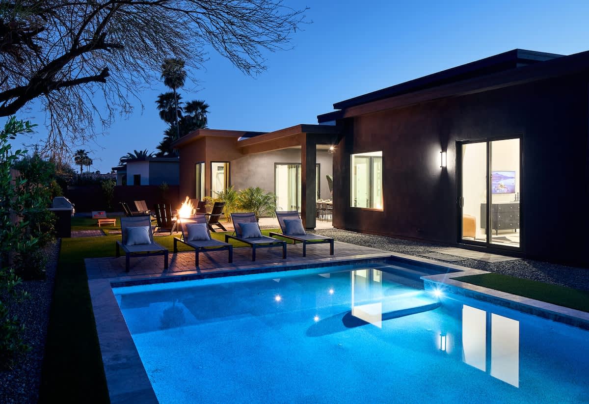 Resort-like backyard with a covered patio offering outdoor dining, a sparkling blue heated pool with sun loungers, yard games, a BBQ grill and a fire pit.