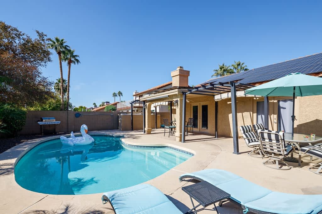 Large private backyard with a sparkling pool, diving board, covered patio with outdoor dining area, sun loungers, and outdoor fire place.
