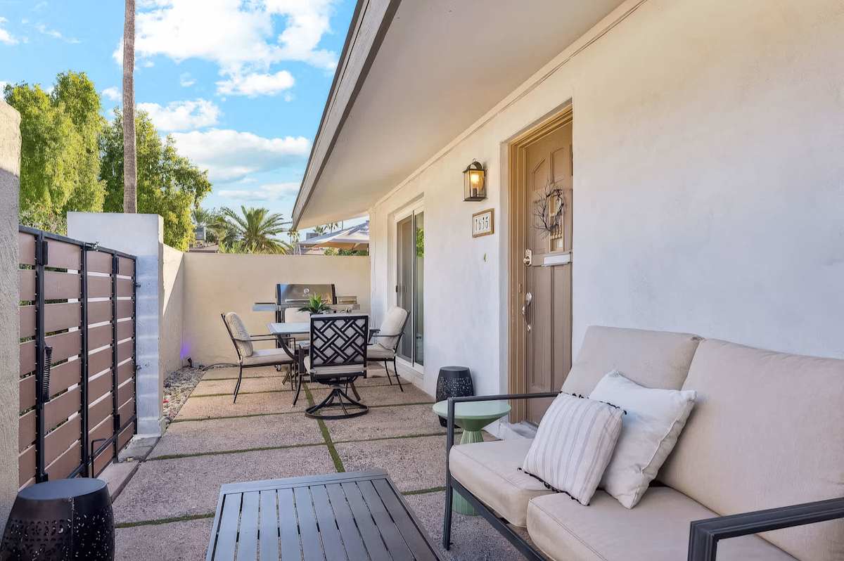 Private patio with lounge seating, a four-seat dining set, gas grill, retractable umbrella, and gated entry framed by swaying palms and Arizona skies.