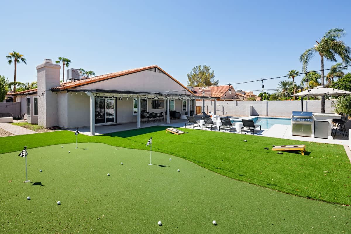 Backyard with putting green, corn hole, BBQ, bar seating, pool, jacuzzi, loungers, outdoor dining table and TV.