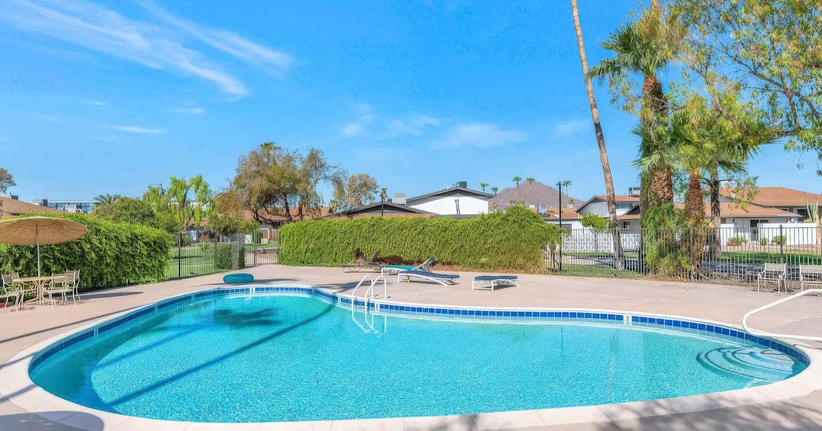 The community pool shimmers under endless Arizona blue skies, ringed by palm trees, lounge chairs, and mountain views just beyond the hedgerow.