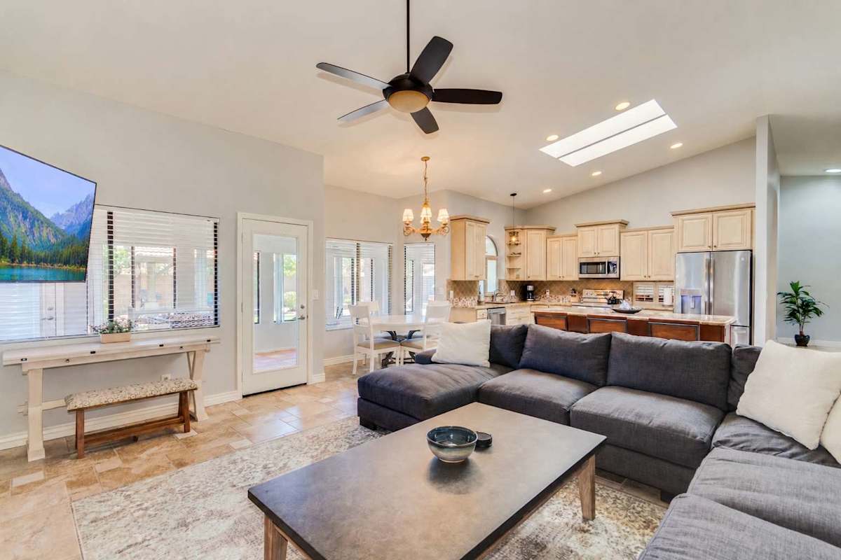 Bright and open floor plan with a large living area that includes a cozy sectional sofa, smart TV, vaulted ceilings, ceiling fans, and direct access to sunroom and kitchen.