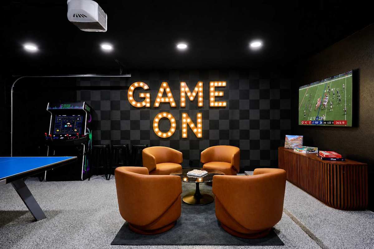 Game room and home gym packed with indoor entertainment, featuring a Peloton bike, ping pong table, arcade game, darts, TV, lounge seating, and a wall-mounted gym system.