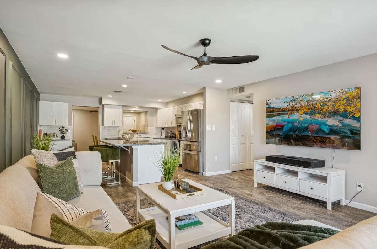 The living room flows seamlessly from the kitchen, anchored by a bold sage green accent wall, plush sectional, large Smart TV, and sliding glass doors opening to the private patio.